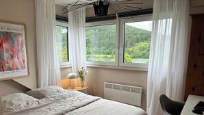 Un dormitorio con cama, escritorio, silla, radiador y ventana con vistas a un bosque y un lago.