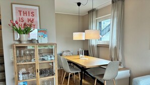Un comedor con mesa de madera, sillas grises, un aparador de vidrio con copas de vino, una lámpara colgante y una ventana con cortinas.