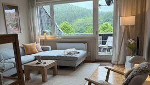 Una sala de estar moderna con sofá, sillón y mesa de centro, ventanas amplias con vista a un bosque y una lámpara.