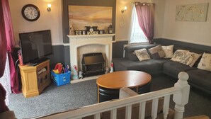A living room with a fireplace, a sofa, a television, and a staircase railing. - Wooden 3-bed Lodge, Sleeps 9/10 With Front Decking (Fleetwood)