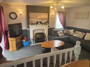A living room with a fireplace, a sofa, a television, and a staircase railing. - Wooden 3-bed Lodge, Sleeps 9/10 With Front Decking (Fleetwood)