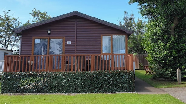 A single-story wooden cabin with a fenced deck, a small porch, and a well-maintained lawn. - Wooden 3-bed Lodge, Sleeps 9/10 With Front Decking (Fleetwood)