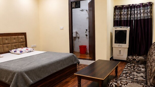 A bedroom with a bed, a wooden table, a sofa, a ceiling fan, and a bathroom with a shower. - OBEROI RESIDENCY  (Pachmarhi)