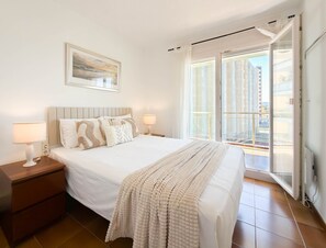 A bedroom with a bed, two bedside tables, a window with a view of buildings, and a painting on the wall. - Maricel apartment with pool and sea views (L'Escala)