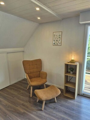 A room with a brown upholstered chair, a matching ottoman, a small wooden shelf with a plant, and a framed picture on the wall. - The edge of the vineyard - the balcony (Andlau)