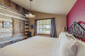 A bedroom with a wooden wall, a bed with white linens, a metal headboard, and a window with curtains.