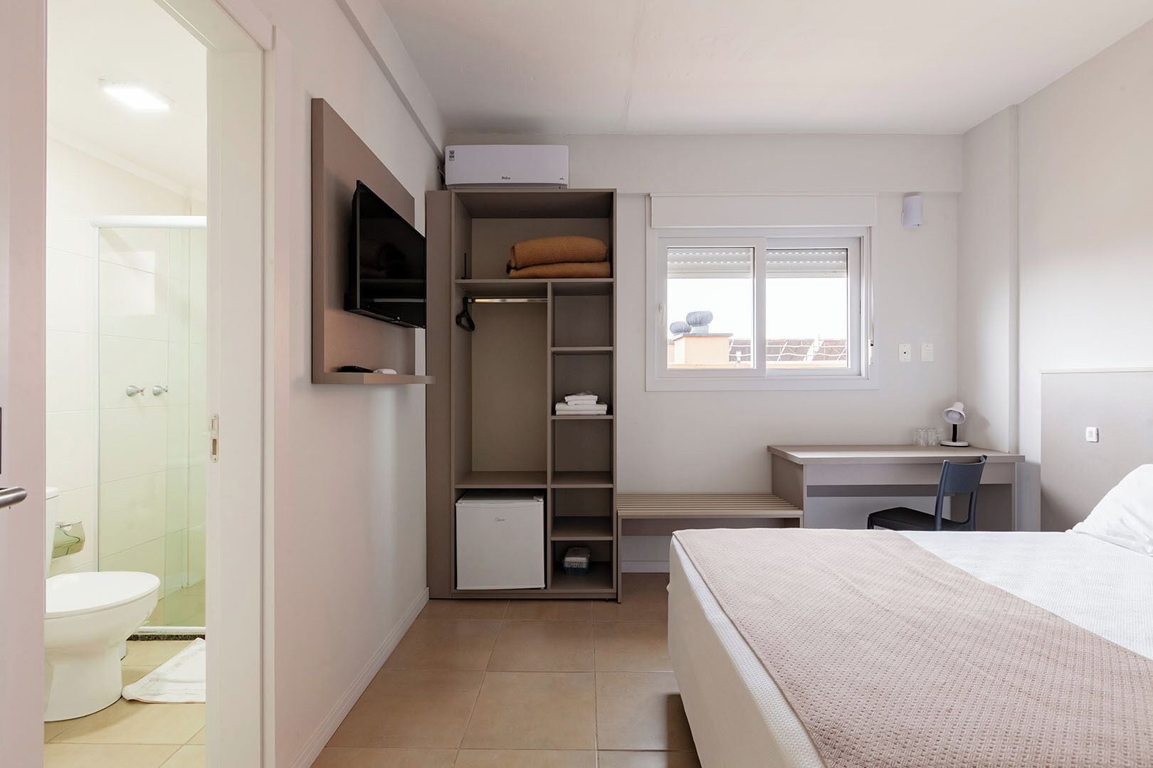A compact hotel room with a bed, a desk, a TV, a wardrobe, and a shower.