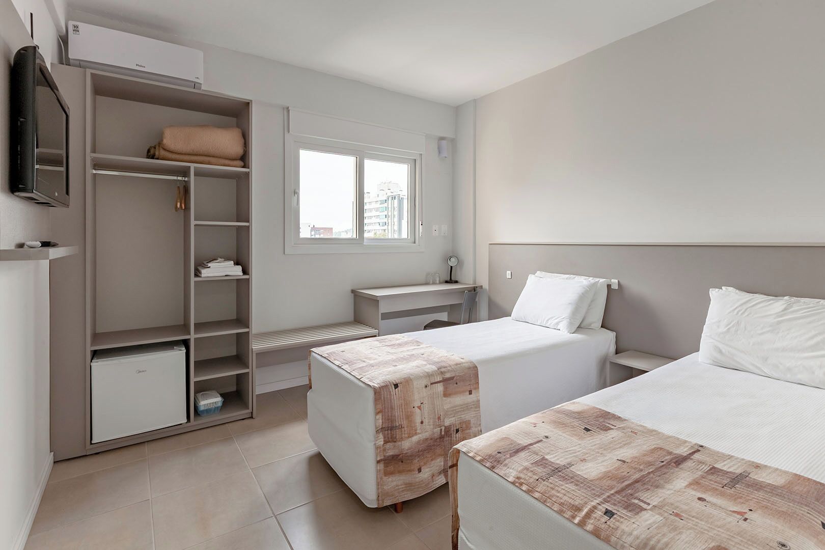 Quarto de hotel com duas camas, um guarda-roupa, uma escrivaninha, uma televisão e uma janela.