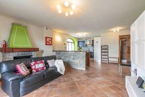 A living room with a fireplace, a stone accent wall, a black leather sofa, and a kitchen in the background. - La Casa Di Olaf With Garden (Bardonecchia)