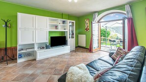 A living room with a TV, white entertainment unit, green walls, a leather sofa, and a view through a glass door. - La Casa Di Olaf With Garden (Bardonecchia)