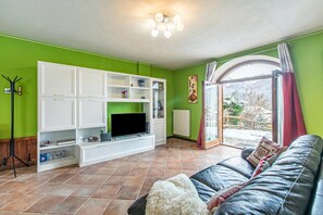 A living room with a green wall, white cabinets, a flat-screen TV, a leather sofa, and a view of a snowy landscape through a glass door. - La Casa Di Olaf With Garden (Bardonecchia)
