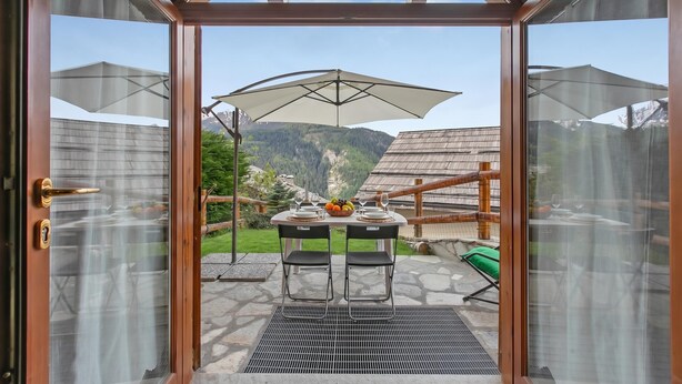 A glass door leading to an outdoor dining area with a table set for two, an umbrella, and a mountainous backdrop. - La Casa Di Olaf With Garden (Bardonecchia)