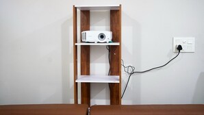 Un projecteur blanc, posé sur une étagère en bois, est adossé à un mur blanc et branché à une prise de courant.