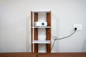 Un projecteur blanc, posé sur une étagère en bois, est adossé à un mur blanc et branché à une prise de courant.