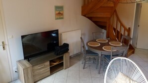 A living room with a TV, wooden staircase, dining table with chairs, and a white wicker chair. - Le50300 Gîte for 5 pers in Avranches, close to Mont Michel ! (Avranches)