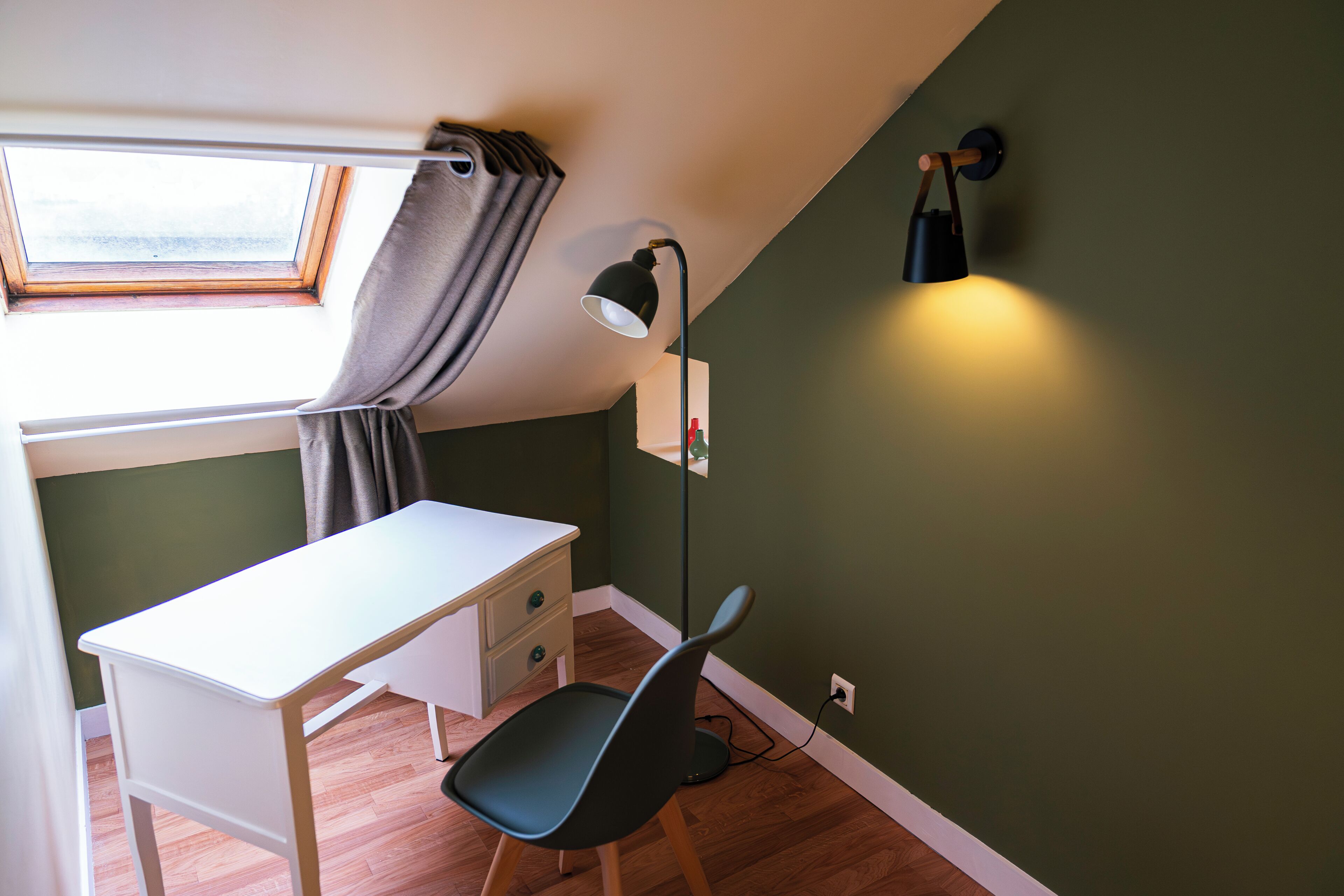 Quarto com escrivaninha, cadeira, janela com cortinas, luminária de chão e espelho fixado na parede.