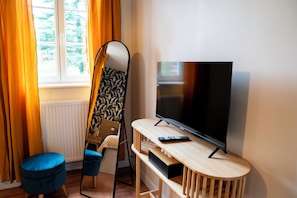 A room with a TV, a blue ottoman, a mirror, and a window with curtains. - Le50300 Gîte for 5 pers in Avranches, close to Mont Michel ! (Avranches)