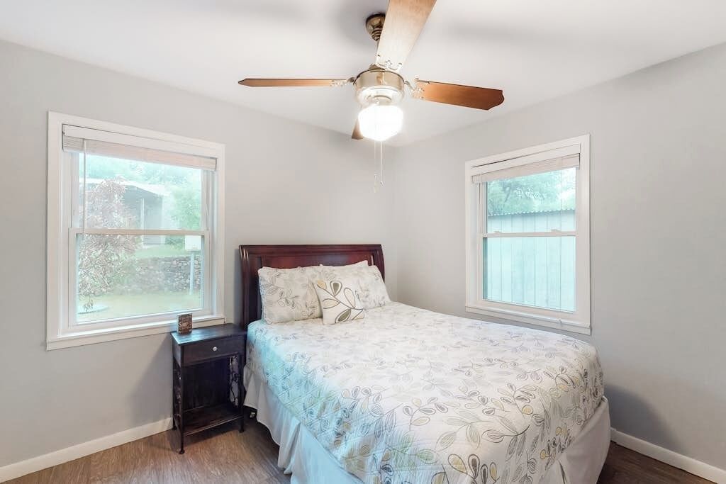A bedroom with a bed, a nightstand, a ceiling fan, and two windows.