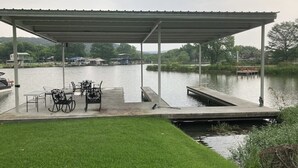 En överbyggd brygga med stolar och ett bord, en gräsyta och en lugn sjö med båtar i bakgrunden.