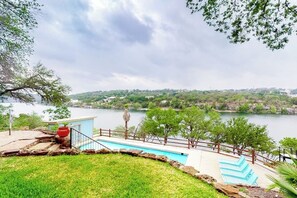A swimming pool with a view of a lake and hills, surrounded by a stone deck and a wooden fence.