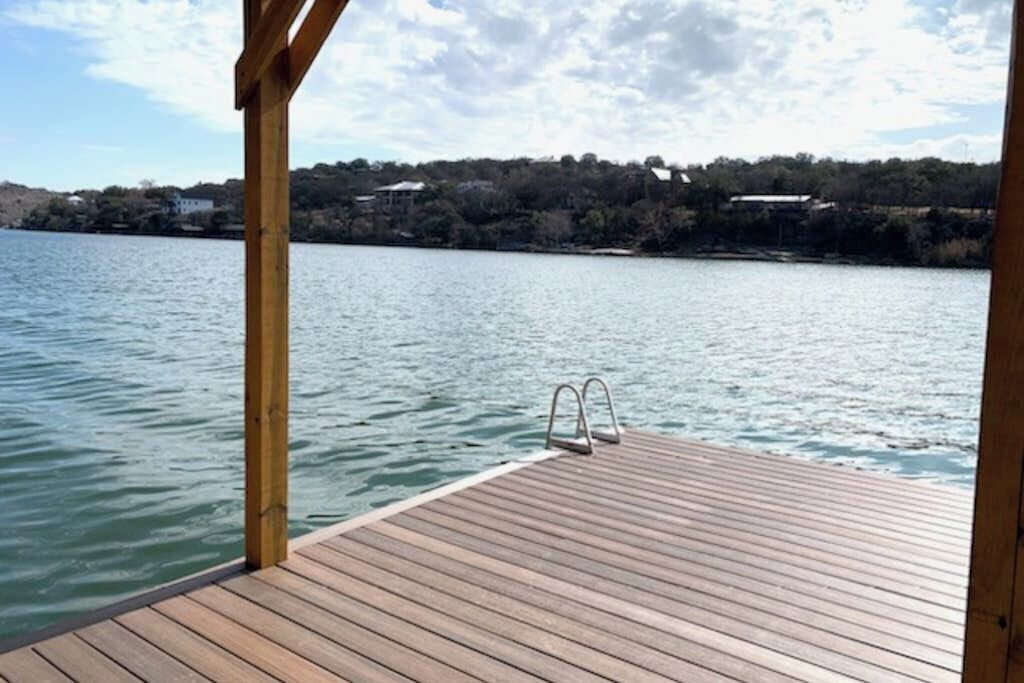 A wooden dock extending into calm water with a metal ladder, surrounded by a clear sky and distant buildings.