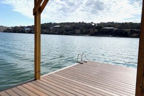 A wooden dock extending into calm water with a metal ladder, surrounded by a clear sky and distant buildings.