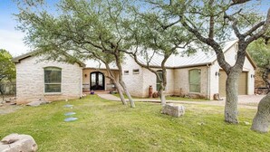A single-story house with a stone facade, a front door, and a small front yard with a tree. - Cliffside Oasis | Deck + Dock | Shuffleboard (Spicewood)