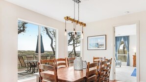 A dining area with a wooden table and chairs, a hanging light fixture, and large windows with a view of trees and a building. - Cliffside Oasis | Deck + Dock | Shuffleboard (Spicewood)