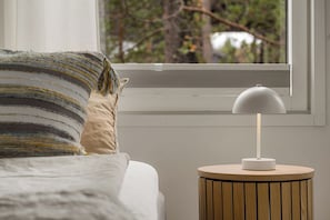 Un lit avec un oreiller à rayures et une table de chevet ronde en bois surmontée d’une lampe blanche.