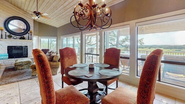A dining area with a round table, four patterned chairs, a chandelier, and large windows offering a view.