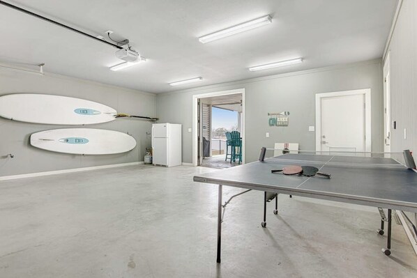 A spacious room with a ping pong table, two surfboards, a refrigerator, and a door leading to an outdoor area. - Lakefront house w/pool, kayaks, ping-pong, gas grill, & paddle boards (Kingsland)