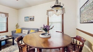 A dining room with a round wooden table, chairs, a chandelier, a sofa with cushions, and a painting on the wall. - Cozy & Cute Cabin with Fenced Patio, Grill, AC & WiFi - Walkable Locale (Fredericksburg)