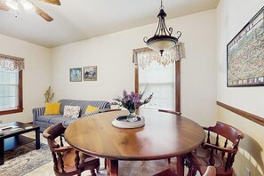 A dining room with a round wooden table, chairs, a chandelier, a sofa with cushions, and a painting on the wall. - Cozy & Cute Cabin with Fenced Patio, Grill, AC & WiFi - Walkable Locale (Fredericksburg)