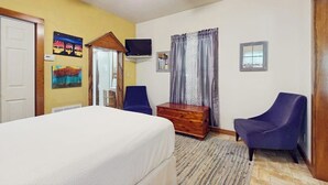 A bedroom with a bed, two chairs, a wooden chest, a television, and a ceiling fan. - Cozy & Cute Cabin with Fenced Patio, Grill, AC & WiFi - Walkable Locale (Fredericksburg)