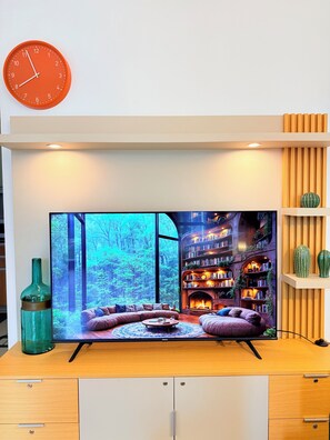 Ein Flachbildfernseher auf einem Holztisch mit Bücherregal, einer Uhr und einem großen Fenster mit Blick auf einen Wald.