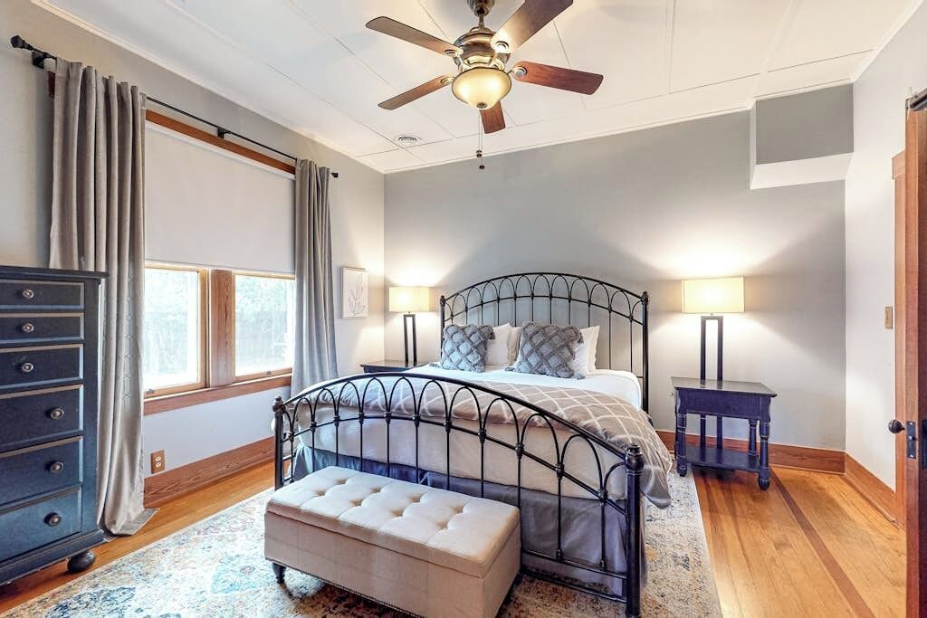 Un dormitorio con cama, ventilador de techo, mesita de noche, cómoda y ventana con cortinas.