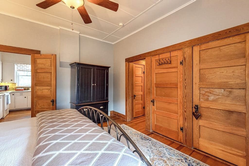 A bedroom with a bed, a ceiling fan, wooden wardrobe, and a kitchen area visible through an open door.