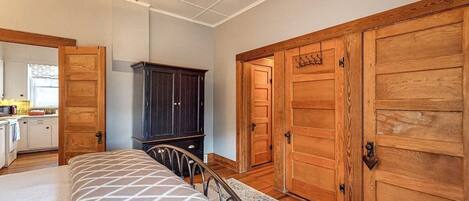 A bedroom with a bed, a ceiling fan, wooden wardrobe, and a kitchen area visible through an open door.