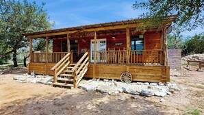 A rustic wooden cabin with a porch, a wheelbarrow, and a sign that reads 'RANCH.' - Dog-friendly gem on 11 acres w/valley views, deck, gas grill, fireplace (Fredericksburg)