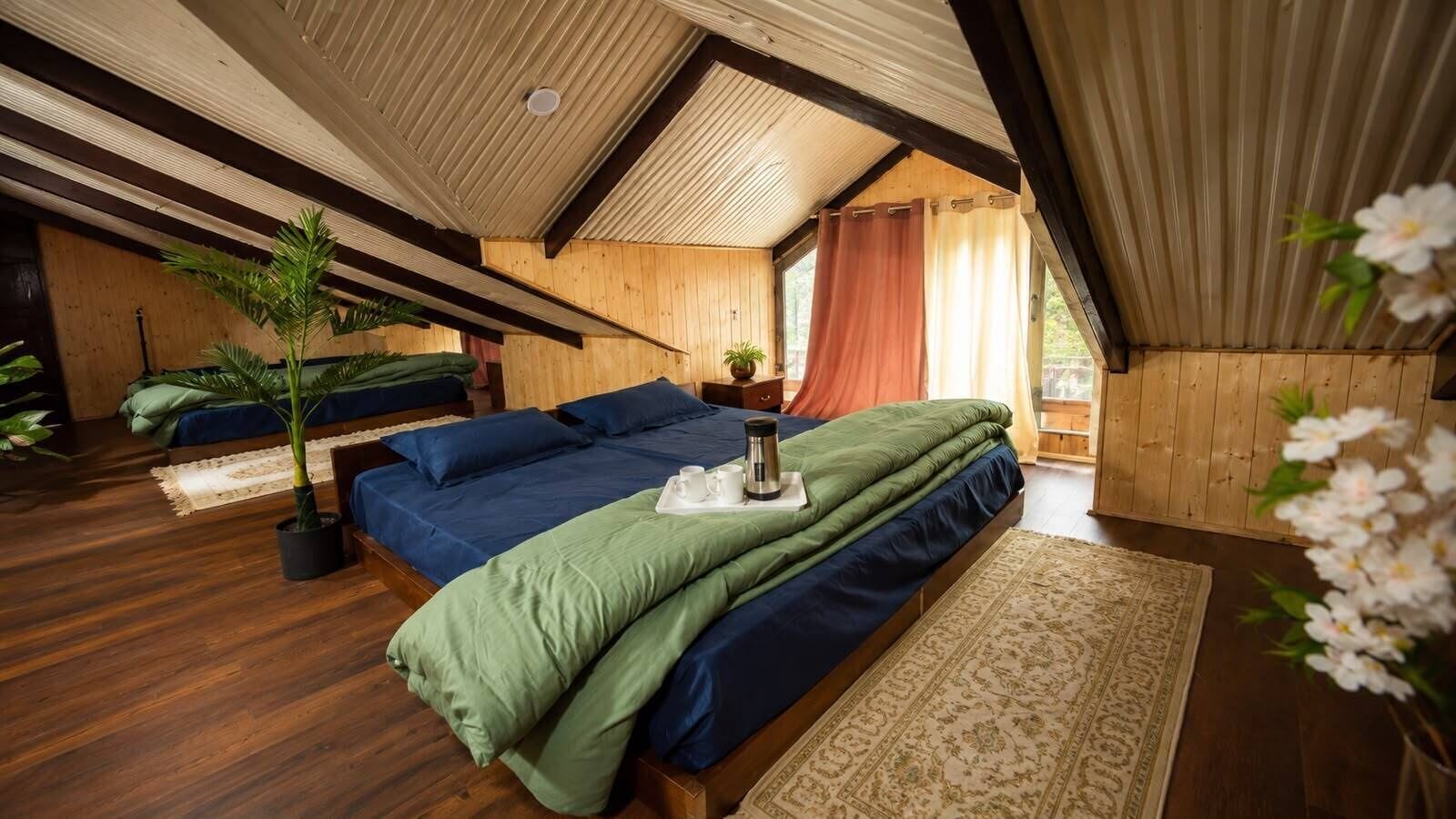 A room with a sloped ceiling, wooden walls, a large bed with blue and green bedding, a potted plant, and a window with curtains.