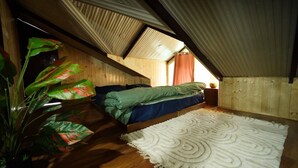 A small, cozy attic bedroom with a sloped ceiling, a single bed, a large plant, and a patterned rug. - The Alpine Hut (Manali)