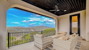 Ein Balkon mit Korbmöbeln, einem Deckenventilator und Blick auf ferne Hügel.