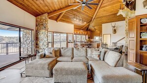 A spacious living room with a large sectional sofa, a ceiling fan, stone walls, and a view through large windows.