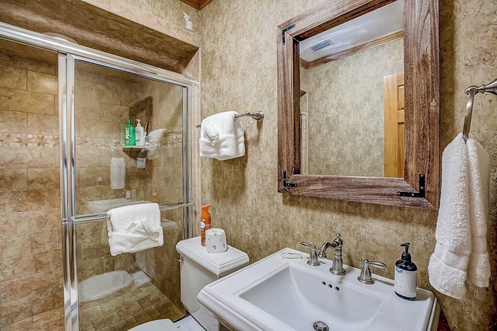 A bathroom with a glass shower enclosure, a white sink with a mirror above it, and a wooden-framed mirror on the wall.