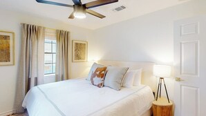 Una habitación de hotel con cama, ventilador de techo, lámpara de noche y ventana con cortinas.