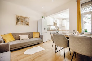 Ein modernes Wohnzimmer mit einer grauen Couch, einem Esstisch mit Stühlen und einer Küche im Hintergrund.