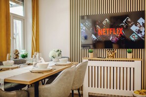 Ein Essbereich mit einem Tisch für vier Personen, einem an der Wand montierten Fernseher, der das Netflix-Logo zeigt, und einem dekorativen Regal mit kleinen Pflanzen.