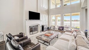 Un salón amplio con chimenea, un televisor de pantalla plana, sofás de cuero y vistas al exterior.