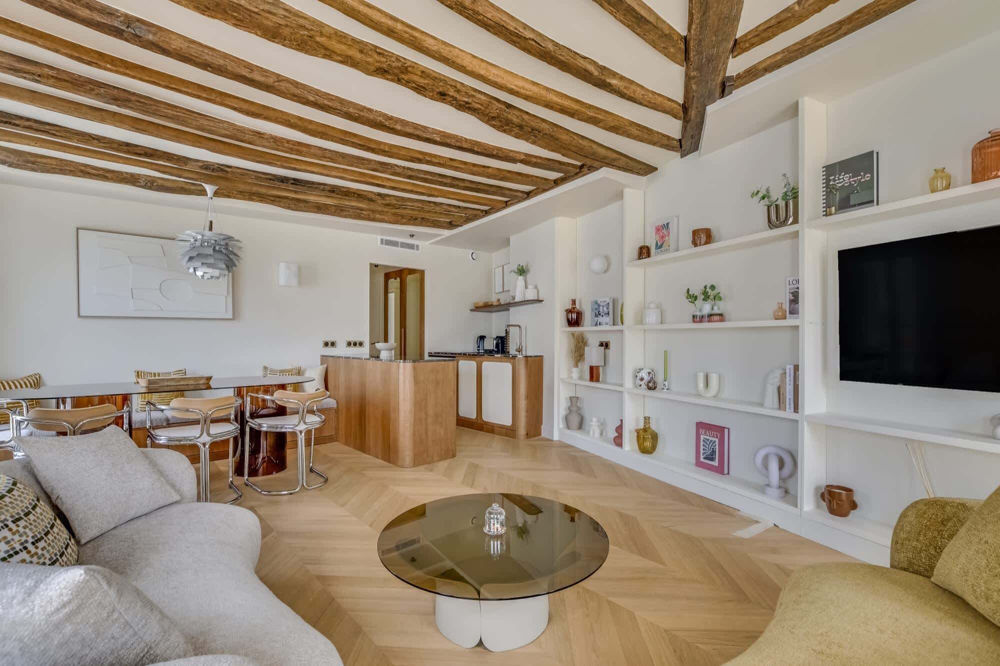 A living room with a wooden ceiling, a glass coffee table, a sofa, a dining area with chairs, and a TV on the wall.
