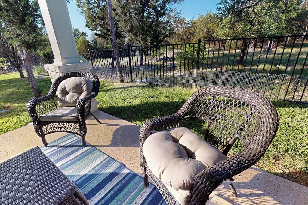 Outdoor wicker furniture with cushions, a striped mat, and a fenced area with trees.